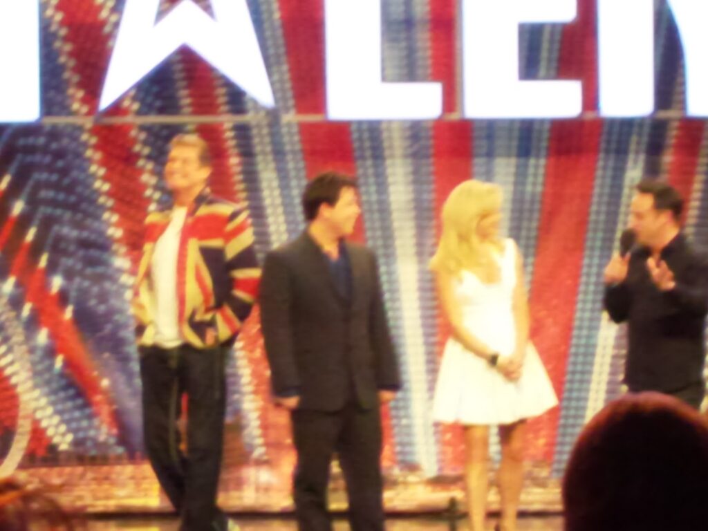 Four people stand on a stage with a colourful, star-patterned backdrop and large white letters. One person wears a Union Jack jacket, another a dark suit, the third a white dress, and the fourth gestures whilst speaking. The image is blurred.