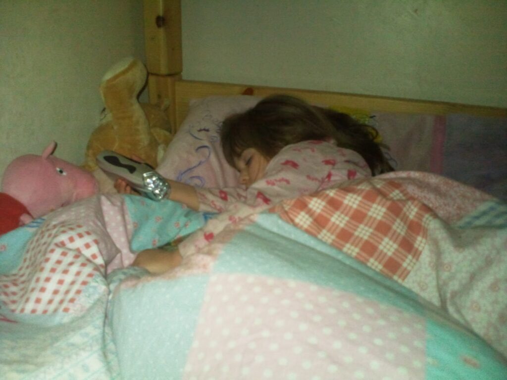 A young girl in patterned pyjamas sleeps in bed, holding a TV remote. She is covered by a colourful patchwork quilt, and soft toys, including a pink Peppa Pig, are beside her. The room looks cosy, with soft lighting and wooden bed frame visible.