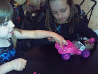 Two young girls sit at a table, playing with a small pink toy car. One girl is holding the toy, while the other is reaching out to touch it. Both girls have light brown hair and are focussed on the toy. The background is softly blurred.