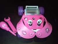 A small pink toy robot with big cartoon eyes, decorated with heart and star stickers. It has purple wheels and two pink tools, a spanner and a bulb-shaped tool, placed next to it on a dark surface.
