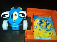 A small toy car made of blue, yellow, white, and black plastic parts sits next to an orange case. The case contains assembly instructions showing the same toy car design with cartoonish eyes on the wheels.