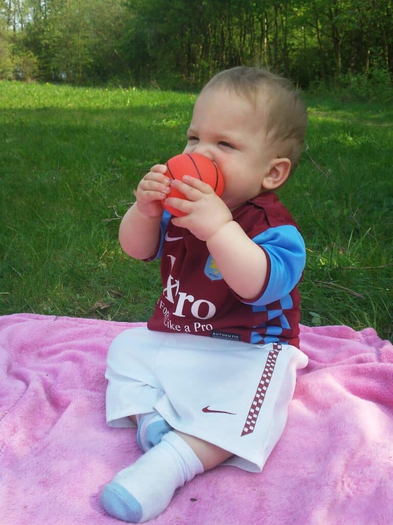 A baby sits on a pink blanket outdoors, wearing a maroon football shirt and white shorts. The baby is holding and chewing on a small orange basketball toy, with trees and green grass visible in the background.