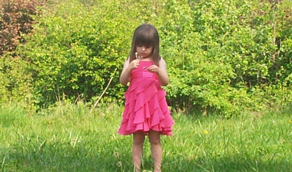 A young girl with straight brown hair stands on the grass wearing a bright pink frilled dress. She looks down at her hands, surrounded by lush, green bushes and trees on a sunny day. The scene feels peaceful and natural.