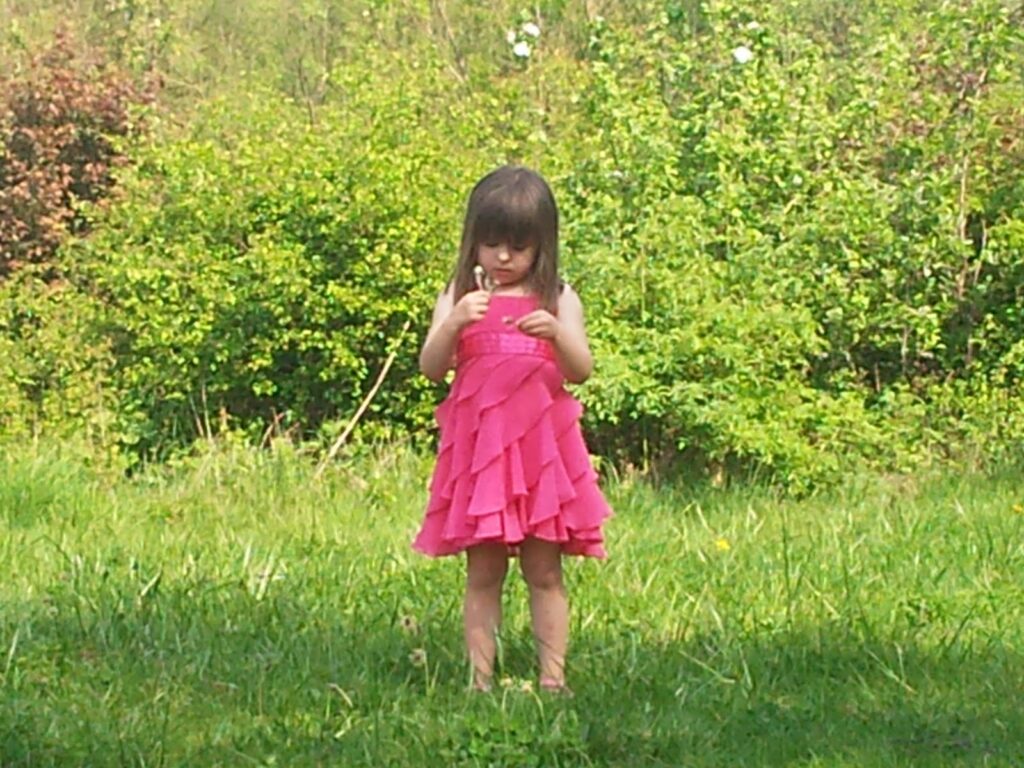 A young child stands barefoot on grass, wearing a bright pink frilled dress. She looks down at something in her hands. Lush green bushes and trees fill the background, and sunlight brightens the outdoor scene.
