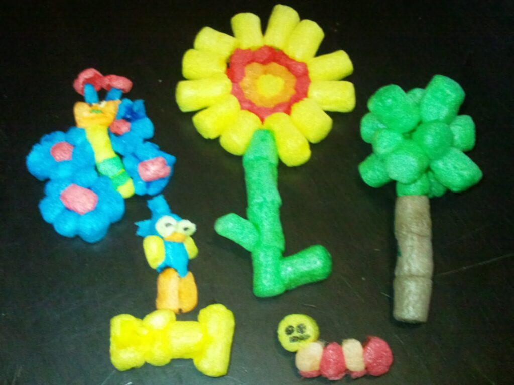 Colourful foam pieces are assembled into four shapes: a butterfly, a tree with a yellow flower, a leafy tree, and a caterpillar. The shapes are arranged side by side on a dark surface, showcasing creative and playful designs.