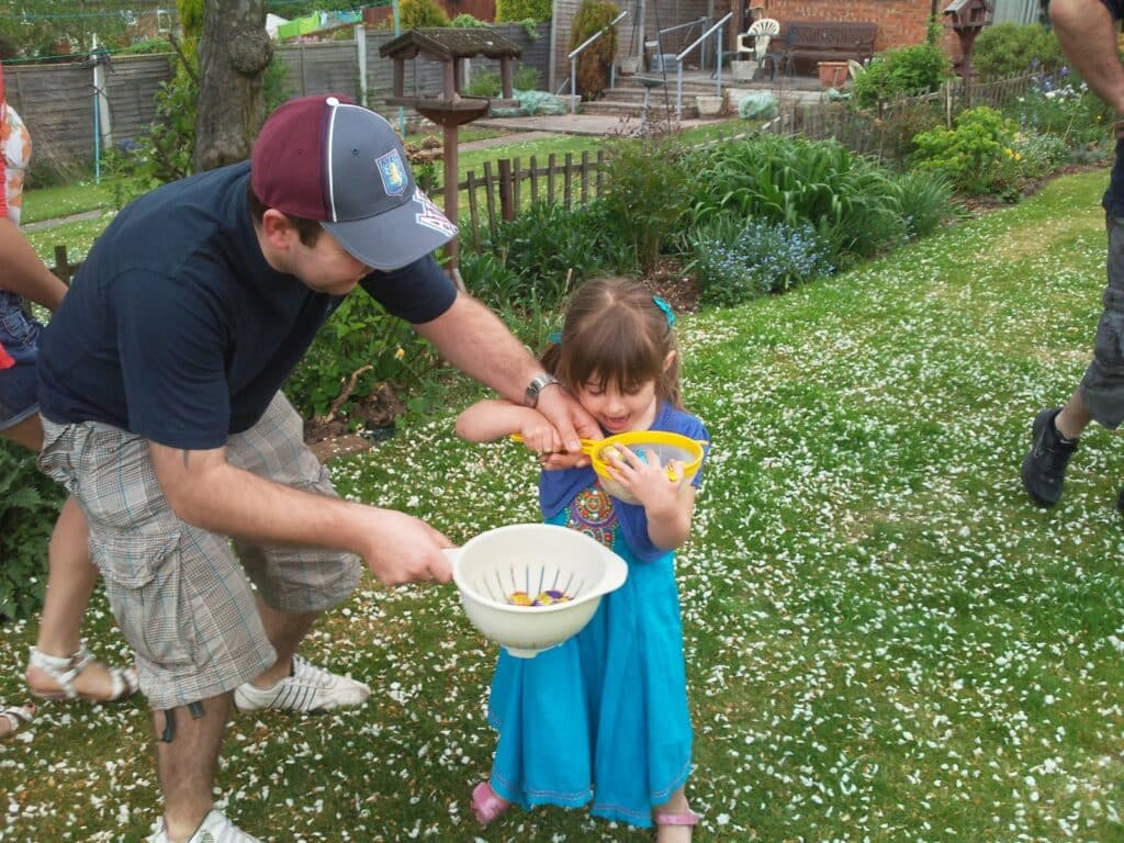 An adult helps a young girl collect Easter eggs in a garden. The girl holds a yellow basket and wears a blue dress. The man, in a cap and checked shorts, holds a white colander with eggs. Flower petals cover the grass; other people walk nearby.