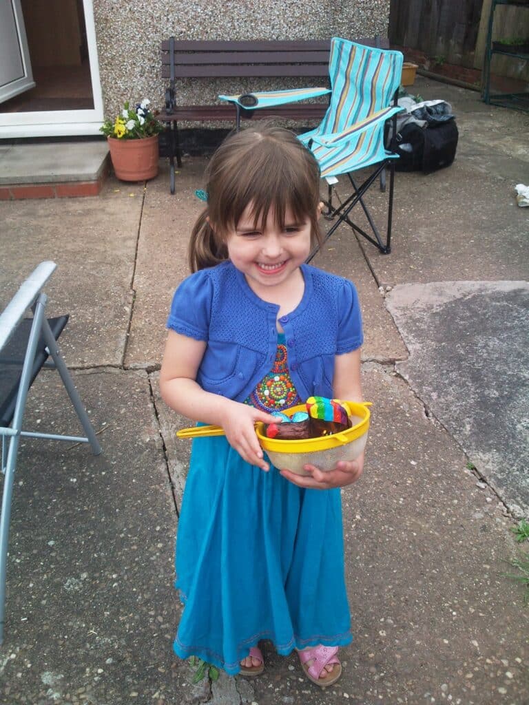 A young girl with brown hair in a ponytail, wearing a blue dress and short cardigan, stands outside on a patio. She is smiling and holding a yellow bowl filled with colourful toys. Folding chairs and a bench are visible in the background.