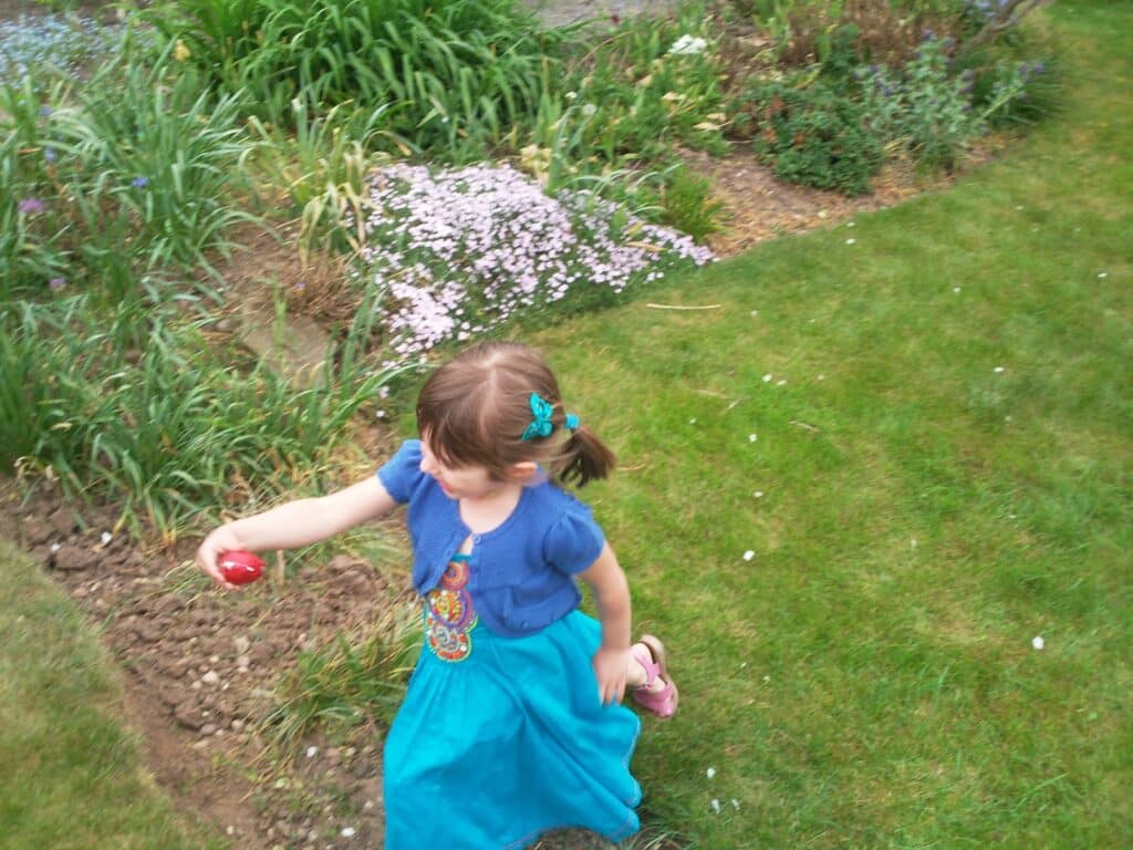 A young girl with brown hair in plaits, wearing a blue dress and matching cardigan, runs on a grassy lawn holding a red egg. She is barefoot, with pink sandals in her other hand. Flowerbeds with green plants and pink flowers are in the background.