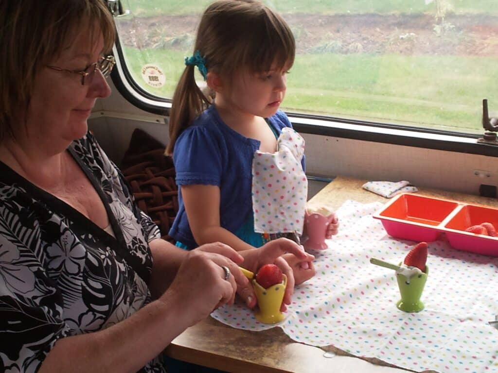 An older woman and a young girl sit at a table in a camper van, preparing strawberries. The woman cuts fruit while the girl watches. Both wear polka-dot serviettes, and colourful cups and containers with strawberries are on the table. Grass is visible outside the window.