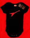 A black baby sleepsuit with short sleeves features a red angular logo resembling a stylised wing or tick across the chest. The sleepsuit is displayed flat against a bright red background with a black tag attached at the collar.