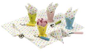 Four pastel-coloured cups (yellow, pink, blue, green) hold polka-dot serviettes folded like ice cream cones. Matching cutlery and spoons are placed on a white cloth with colourful polka dots, creating a playful, ice cream-themed table setting.