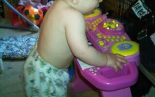 A baby wearing patterned trousers and no shirt stands upright, playing with a pink and yellow toy keyboard. The background is cluttered with shoes, toys, and household items, creating a casual indoor setting. The baby appears focused on the toy.