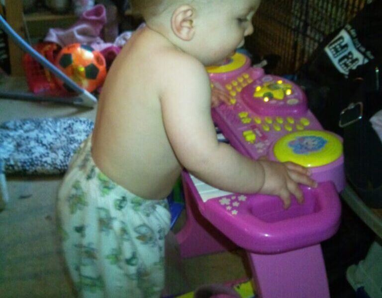 A baby wearing patterned trousers and no shirt stands upright, playing with a pink and yellow toy keyboard. The background is cluttered with shoes, toys, and household items, creating a casual indoor setting. The baby appears focused on the toy.