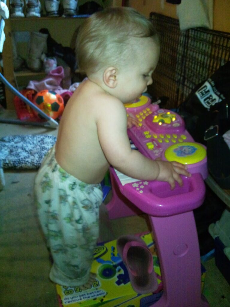 A baby wearing patterned trousers stands and plays with a pink and yellow toy piano. The baby is barefoot and topless, and the room has various items scattered around, including shoes, a football, and boxes in the background.