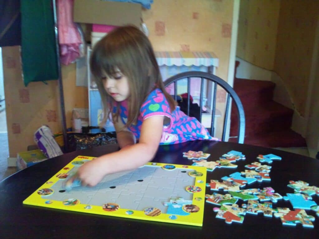 A young girl with straight brown hair, wearing a colourful patterned dress, sits at a black table and works on assembling a jigsaw with cartoon images. Several puzzle pieces are scattered on the table beside the yellow jigsaw frame.
