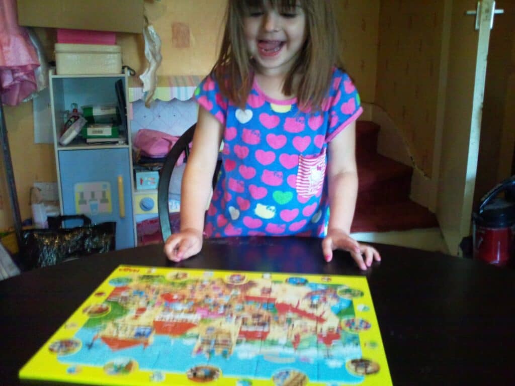 A young girl with brown hair, wearing a blue shirt with pink hearts, smiles while looking at a colourful puzzle on a dark table. The room has shelves, toys, and a staircase in the background, creating a cosy, playful atmosphere.