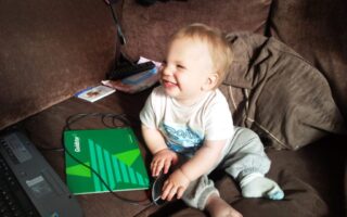 A smiling toddler sits on a brown sofa using a computer mouse. A green notebook labelled "Goldstar," a laptop, and a remote control are nearby. The child wears a white shirt and grey tracksuit bottoms, with sunlight illuminating their face.