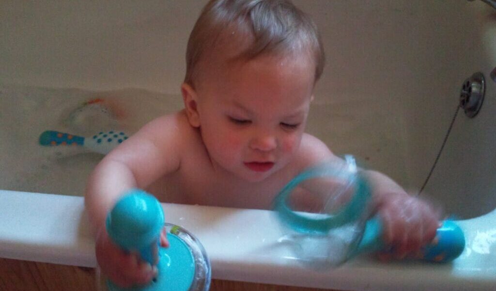 A toddler with light brown hair is sitting in a bath, playing with two large blue bath toys. The child is focused on the toys, with water and more bath toys visible in the background. The bath has a white rim and a wooden panel exterior.