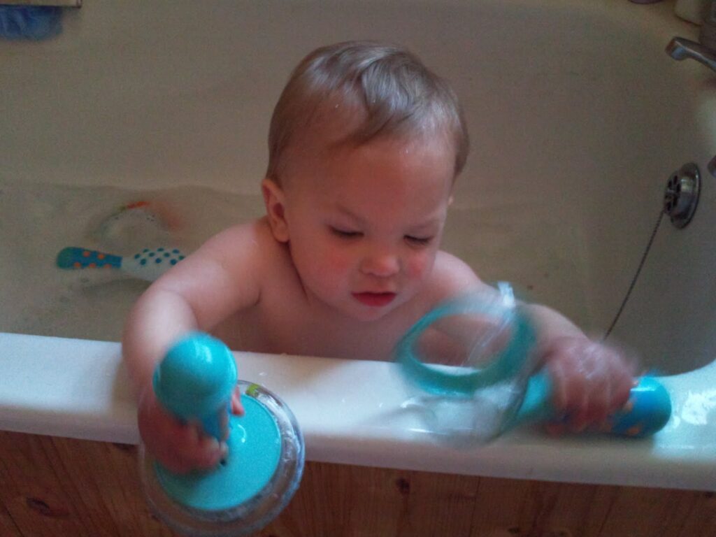 A young child is sitting in a bath, playing with two blue bath toys. The child is reaching over the edge of the bath, holding a toy in each hand, with wet hair and a focused expression. The bath is filled partway with water and other bath toys are visible.