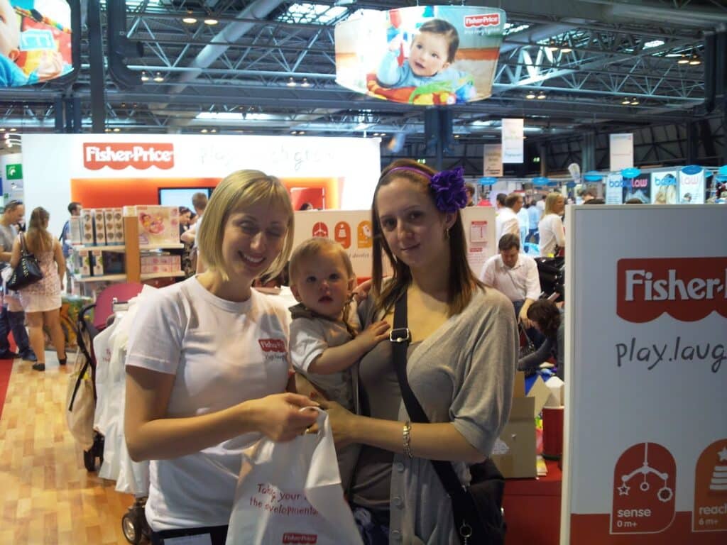 Two women stand together at a Fisher-Price stand. One woman holds a baby, while the other, in a white shirt, holds a tote bag. Colourful displays, pushchairs, and people fill the background. Overhead monitors show a baby advertisement.