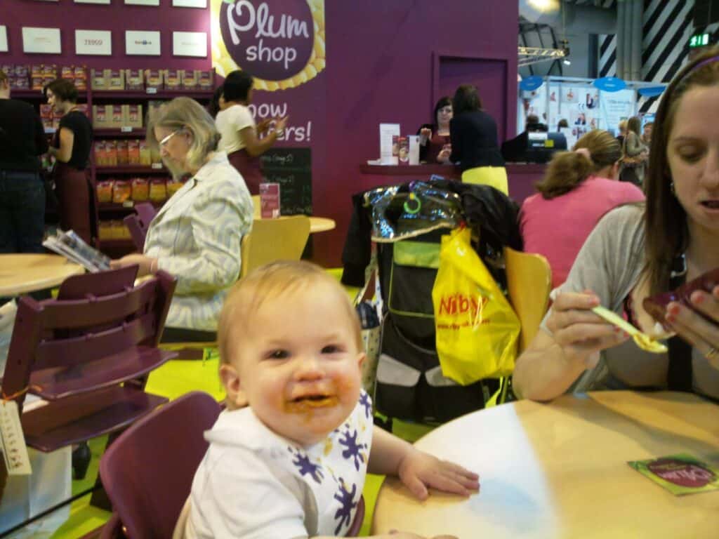 A smiling baby with food smeared on their mouth sits at a table in a busy café. The baby wears a white bib and purple shirt. An adult next to the baby eats from a bowl. In the background, people sit at tables and a purple “Plum shop” wall display is visible.