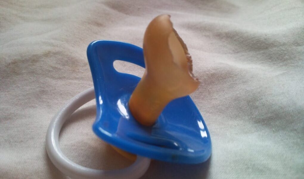 A blue baby dummy with a white ring lies on a light fabric surface. The rubber teat of the dummy is visibly damaged, with a large chunk bitten off, making it unusable.