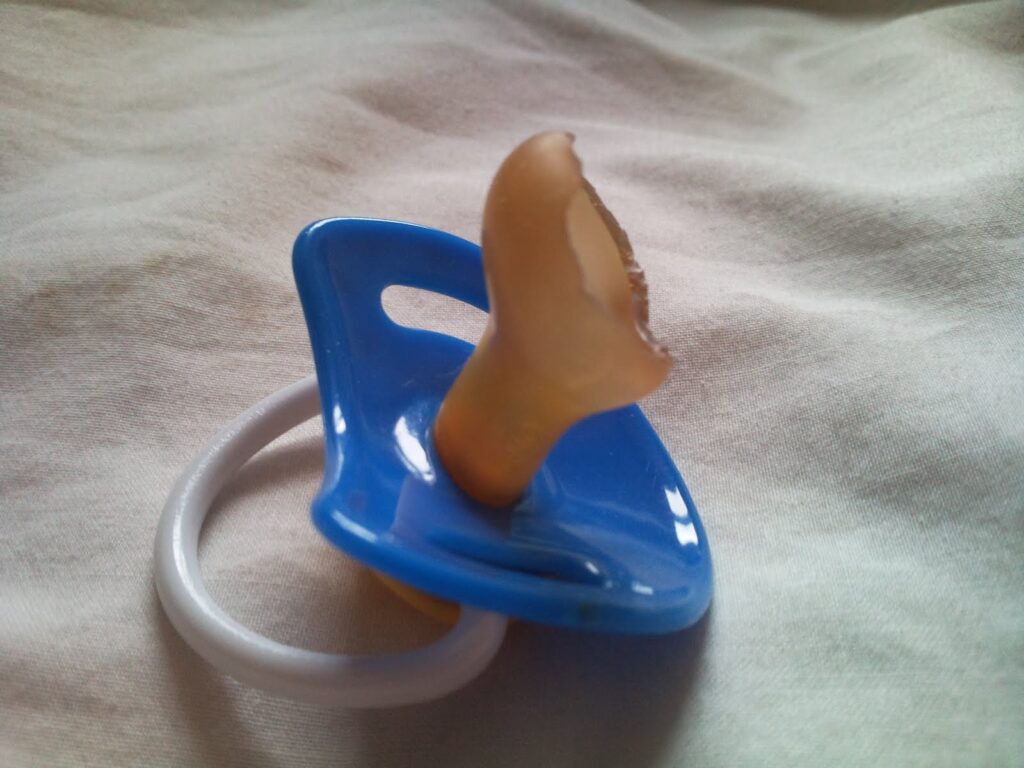 A blue and white baby dummy with a circular handle lies on a soft, light-coloured fabric. The silicone teat is noticeably chewed and damaged, with the top part missing and jagged edges visible.