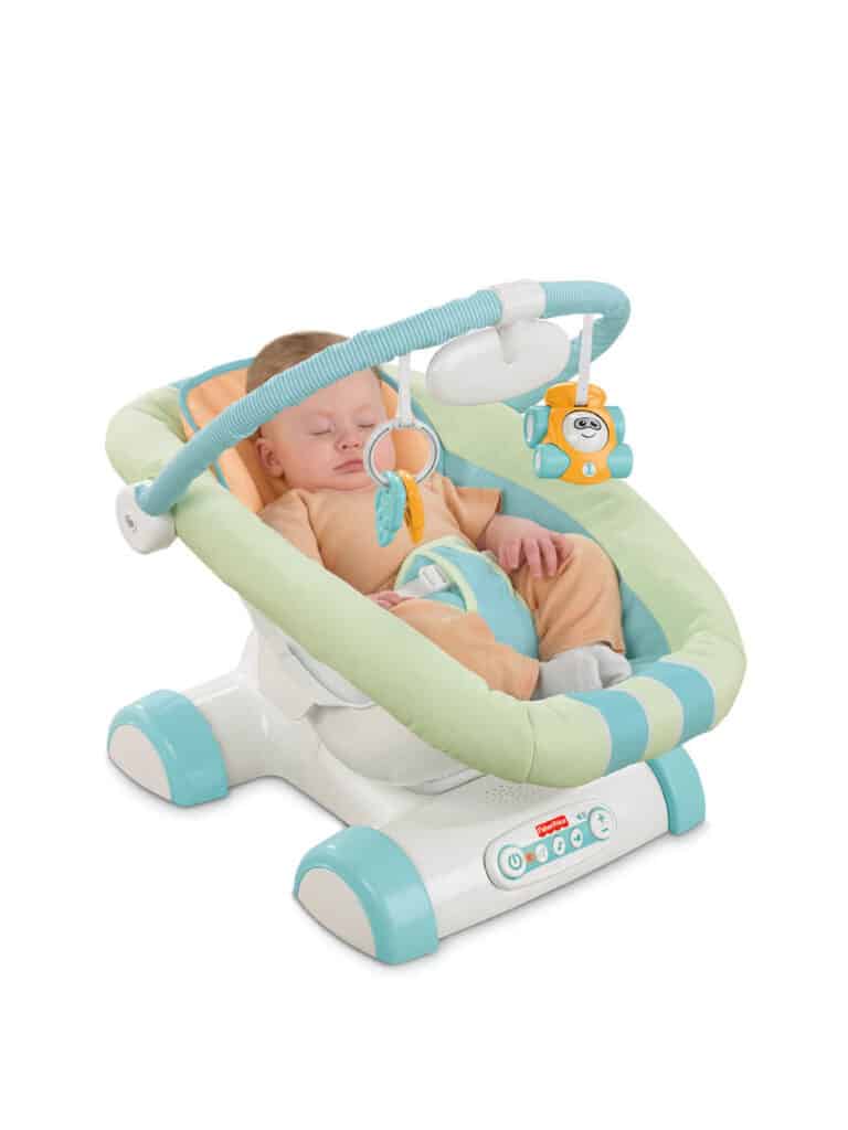 A sleeping baby is secured in a pastel green and turquoise baby rocker with a toy bar. The rocker has a soft cushion and two hanging toys: a ring and a bear. The baby is swaddled in a peach outfit and appears comfortable and peaceful.