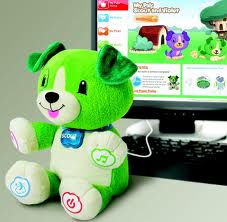 A green and white plush toy dog with a blue collar labelled "Scout" sits in front of a computer monitor displaying a colourful children’s website. The toy has icons on its paws and is connected to the computer with a white cable.
