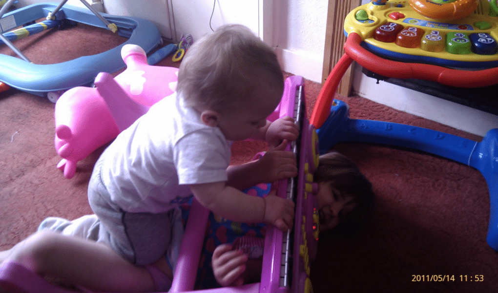 A baby in a light babygro leans over a colourful toy piano, gripping its keys, while an older child lies underneath the toy, partially obscured, with only their face and hands visible. A pink ride-on toy and other play items are in the background on a carpeted floor.