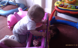 A baby in a light babygro leans over a colourful toy piano, gripping its keys, while an older child lies underneath the toy, partially obscured, with only their face and hands visible. A pink ride-on toy and other play items are in the background on a carpeted floor.