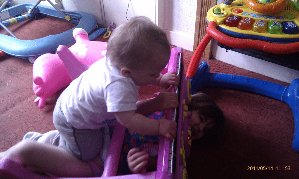 A baby climbs over a toddler lying on the floor behind a pink toy piano. The toddler’s face is partly hidden by the piano. Toys are scattered around on a reddish carpet. Sunlight streams in from the left. A date stamp reads “14/05/2011 11:53”.