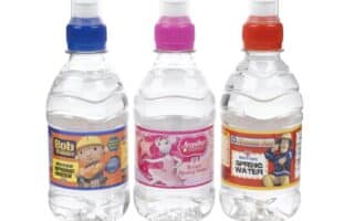 Three clear plastic bottles of water with colourful caps (blue, pink, red) stand side by side. Each features a label with a children’s character: Bob the Builder, Angelino the Pony, and Fireman Sam, with "British Spring Water" text visible.