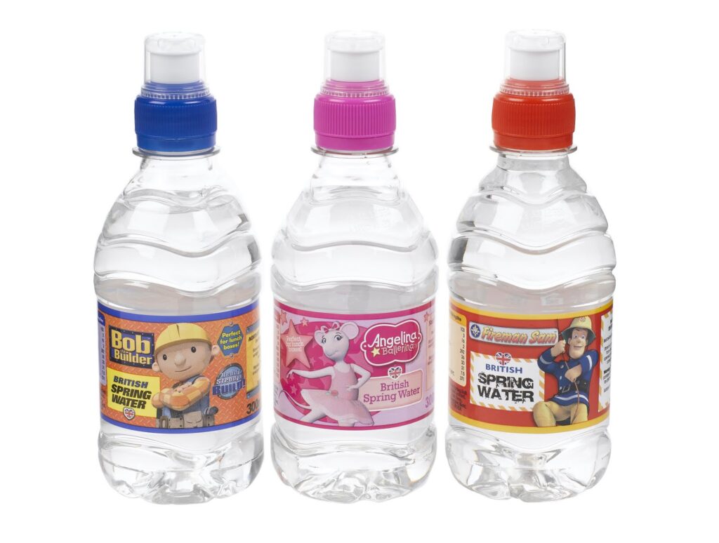 Three plastic water bottles with colourful caps—blue, pink, and red. Each bottle has a character-themed label: a builder, a unicorn, and a superhero, with text "British Spring Water." The bottles are clear and filled with water.
