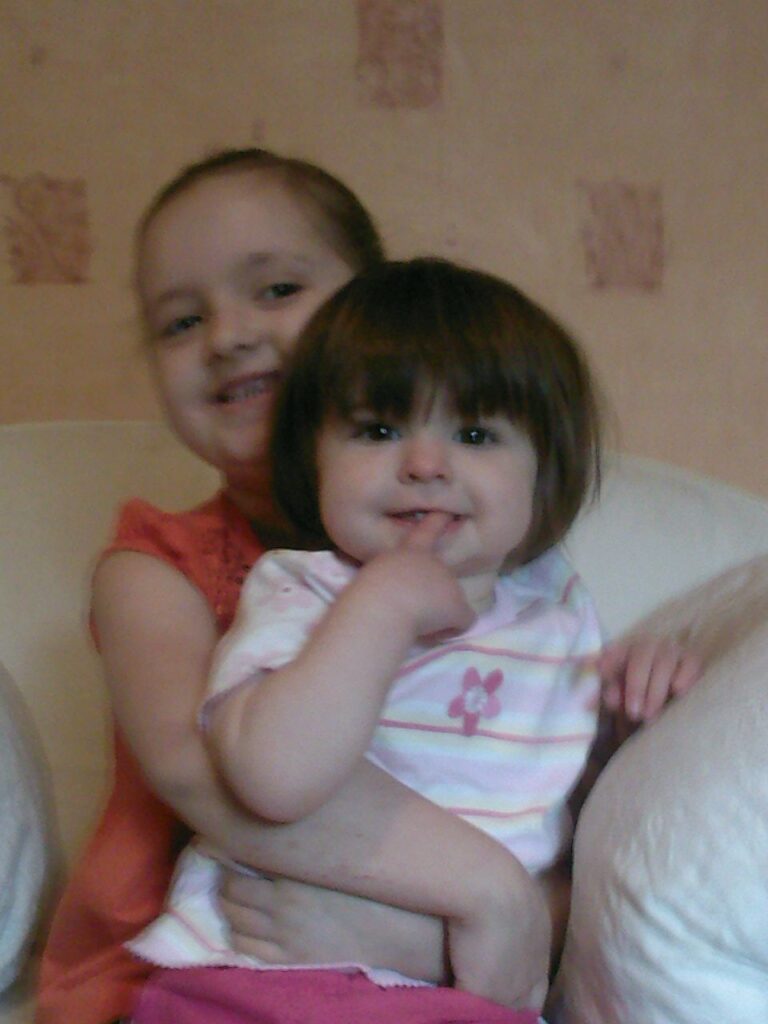 Two young girls sit on a light sofa. The older girl, smiling and wearing a sleeveless coral top, hugs the younger girl from behind. The younger girl has short brown hair and a striped top, and she smiles with a finger near her mouth.