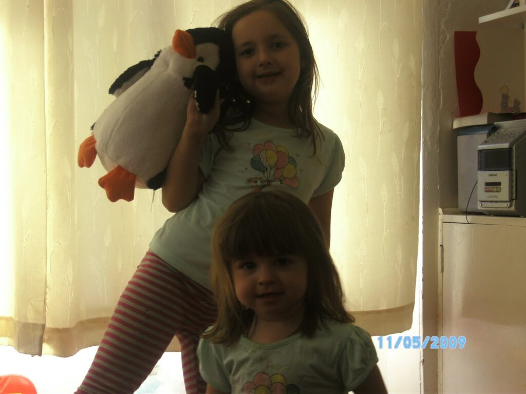 Two young girls stand indoors in front of beige curtains. The older girl holds a stuffed penguin and wears a light blue shirt and striped trousers. The younger girl stands in front, also in a light blue shirt. Sunlight comes from behind them, creating a silhouette effect.