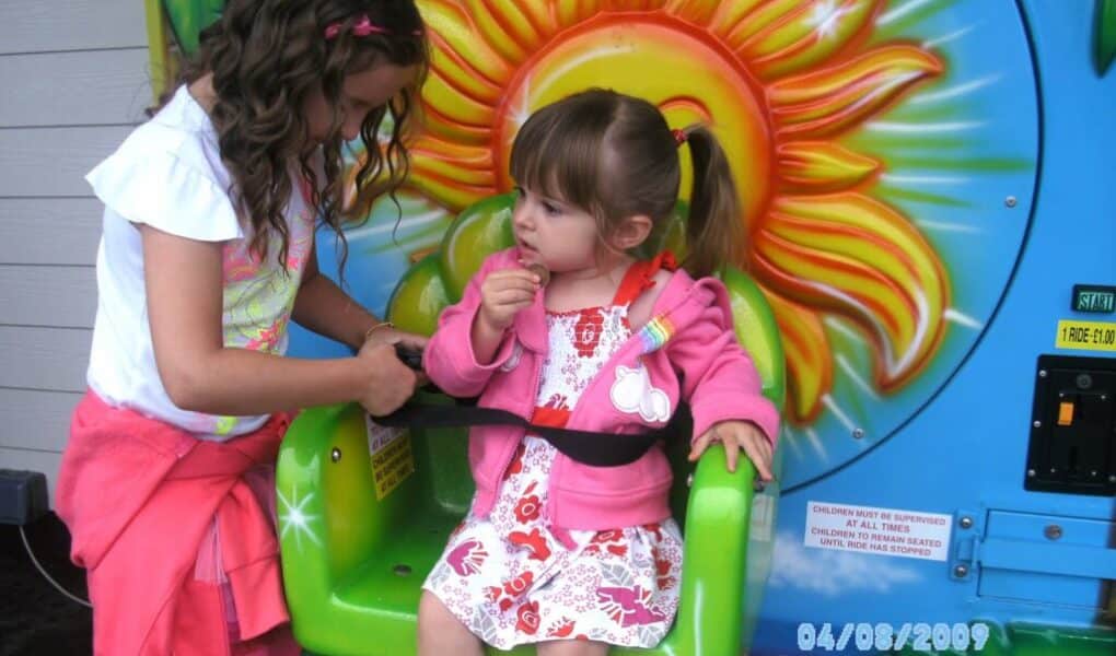 Two young girls are at a funfair ride with a colourful sun design. The older girl, standing, helps fasten the seatbelt for the younger girl, who sits in a green seat wearing a pink jacket and floral dress. The date “08/04/2009” is visible.