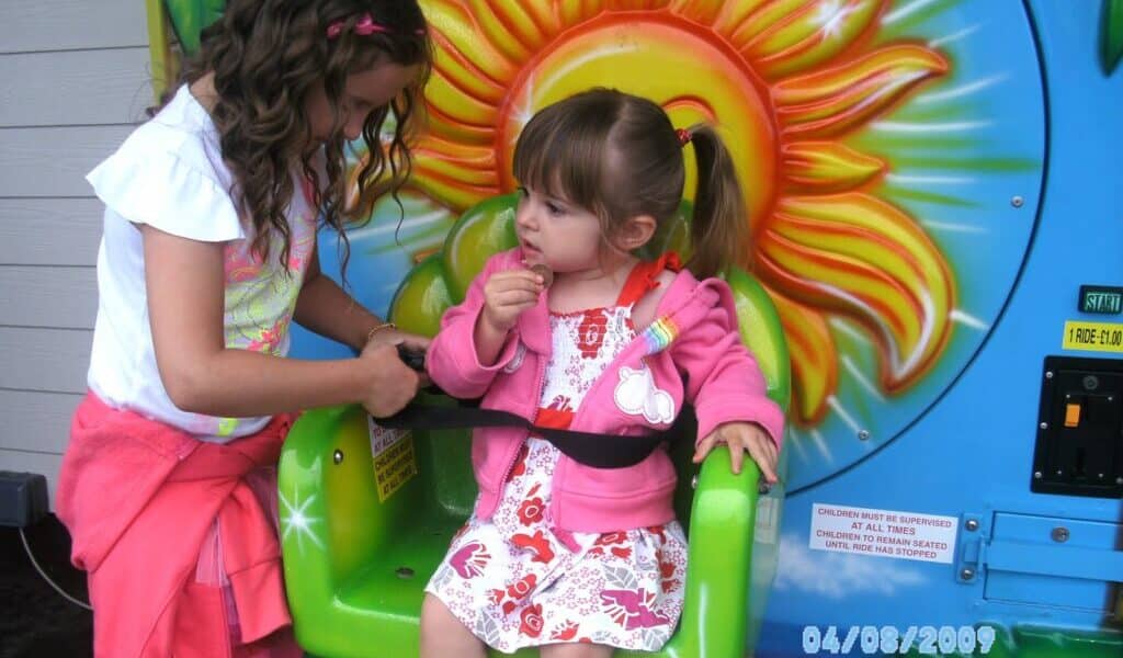 Two young girls are at a funfair ride with a colourful sun design. The older girl, standing, helps fasten the seatbelt for the younger girl, who sits in a green seat wearing a pink jacket and floral dress. The date “08/04/2009” is visible.