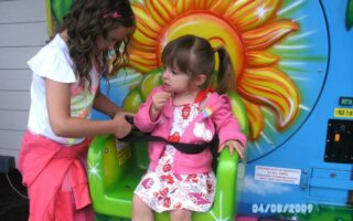 Two young girls are at a funfair ride with a colourful sun design. The older girl, standing, helps fasten the seatbelt for the younger girl, who sits in a green seat wearing a pink jacket and floral dress. The date “08/04/2009” is visible.
