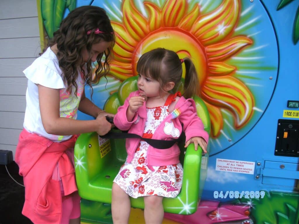 Two young girls are at a colourful amusement ride with a sun design. The older girl helps buckle the younger girl, who is seated wearing a pink jacket and flower dress. The date “04/08/2009” is visible on the bottom right of the image.