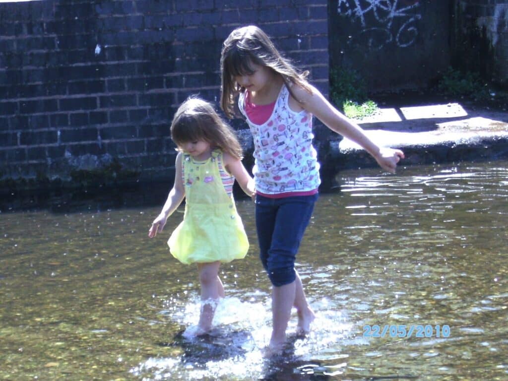 Two young girls wade barefoot in shallow water, holding hands. The older girl wears dark leggings and a patterned vest top, guiding the younger girl in a yellow dress. Sunlight reflects on the water; a brick wall and graffiti are visible in the background.