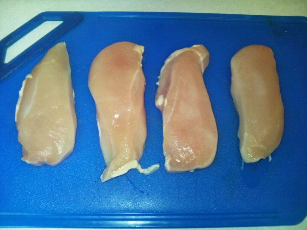 Four raw, skinless chicken breasts are arranged in a row on a bright blue plastic chopping board, with the handle of the board visible on the left side. The chicken pieces are pale pink and unseasoned, ready for preparation.