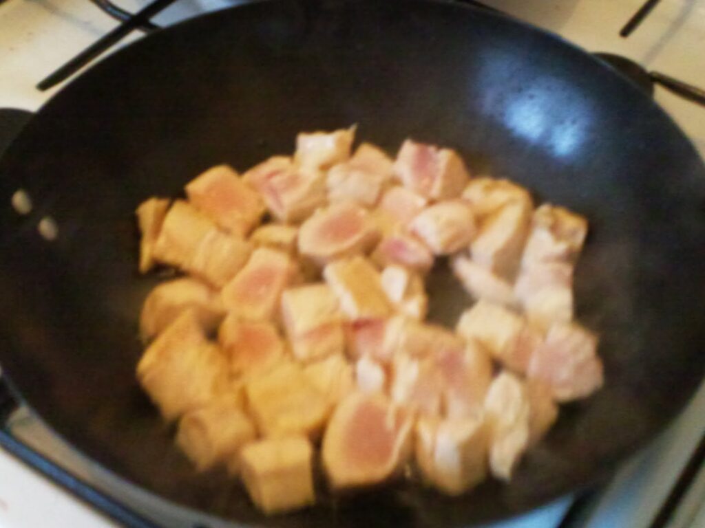 Chunks of raw chicken are cooking in a black frying pan on a hob. The pieces are pink and starting to brown slightly on the edges, indicating the cooking process has just begun. Steam rises from the pan, and the stove grates are visible in the background.