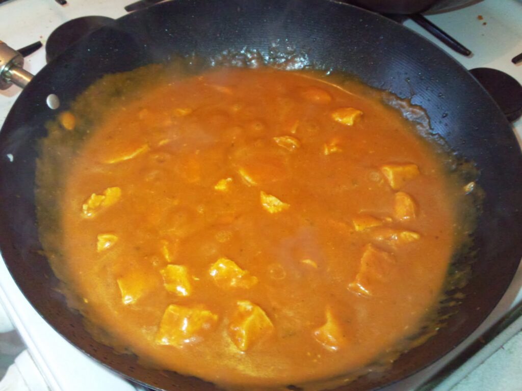 Chunks of chicken simmer in a rich, orange-brown curry sauce inside a black frying pan set on a hob. Steam rises from the hot dish, and the sauce appears creamy and smooth, coating the chicken pieces. The hob burners are visible in the background.