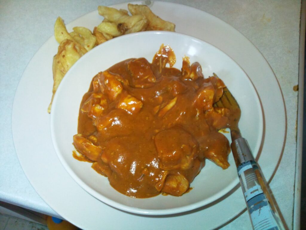 A white bowl filled with chicken pieces in a thick reddish-brown curry sauce sits on a white plate. On the plate next to the bowl are several chunky, golden-brown potato chips. A fork rests in the bowl. The background is a white worktop.