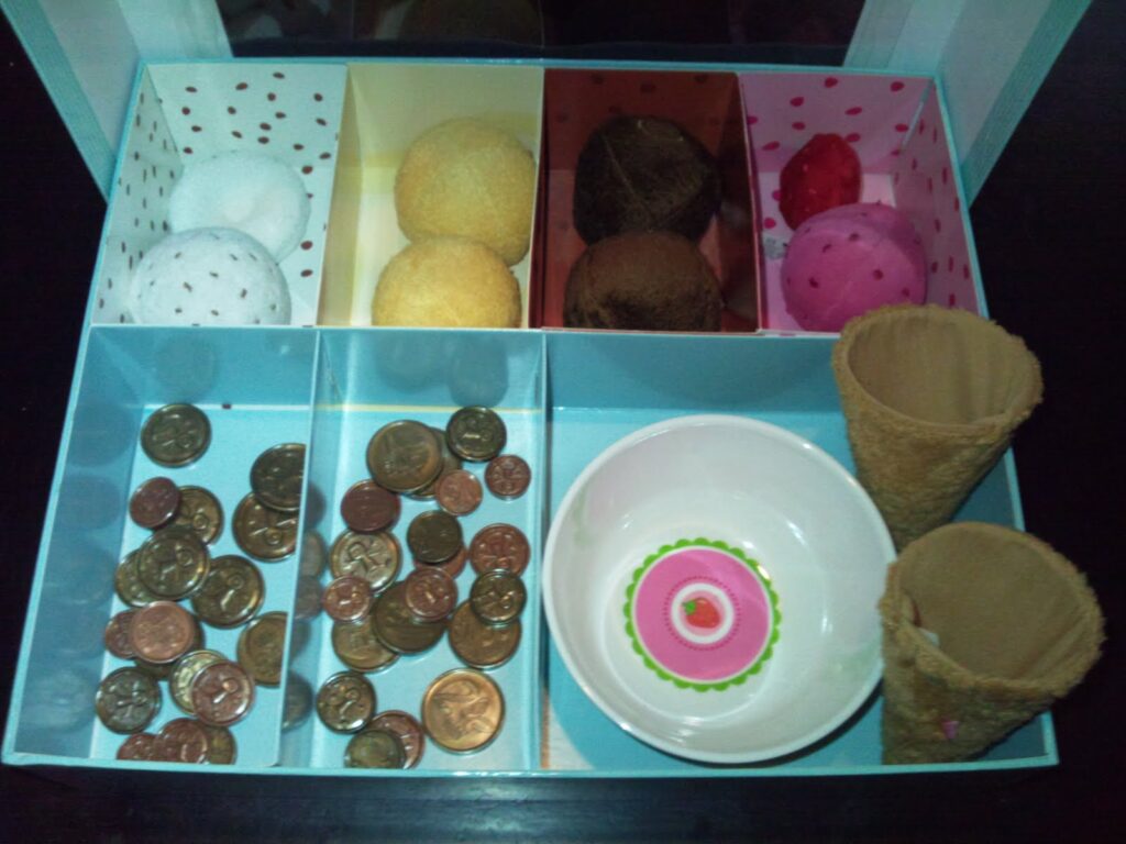 A box with separate compartments holds toy ice cream scoops in various colours, several U.S. coins, two empty waffle cones, and a small, white bowl with a floral pattern at the centre, all arranged neatly.