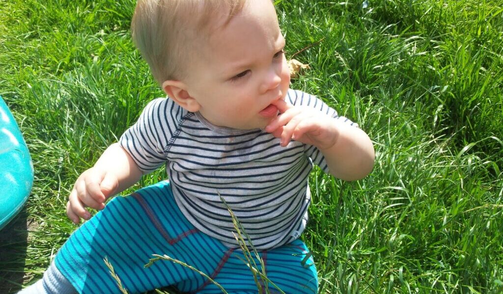 A baby with blonde hair sits in tall green grass, wearing a striped short-sleeve shirt and blue striped trousers. The child touches their mouth with one hand and looks thoughtfully to the side, sunlight highlighting their face and hair.