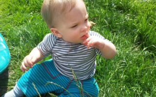 A baby with blonde hair sits in tall green grass, wearing a striped short-sleeve shirt and blue striped trousers. The child touches their mouth with one hand and looks thoughtfully to the side, sunlight highlighting their face and hair.