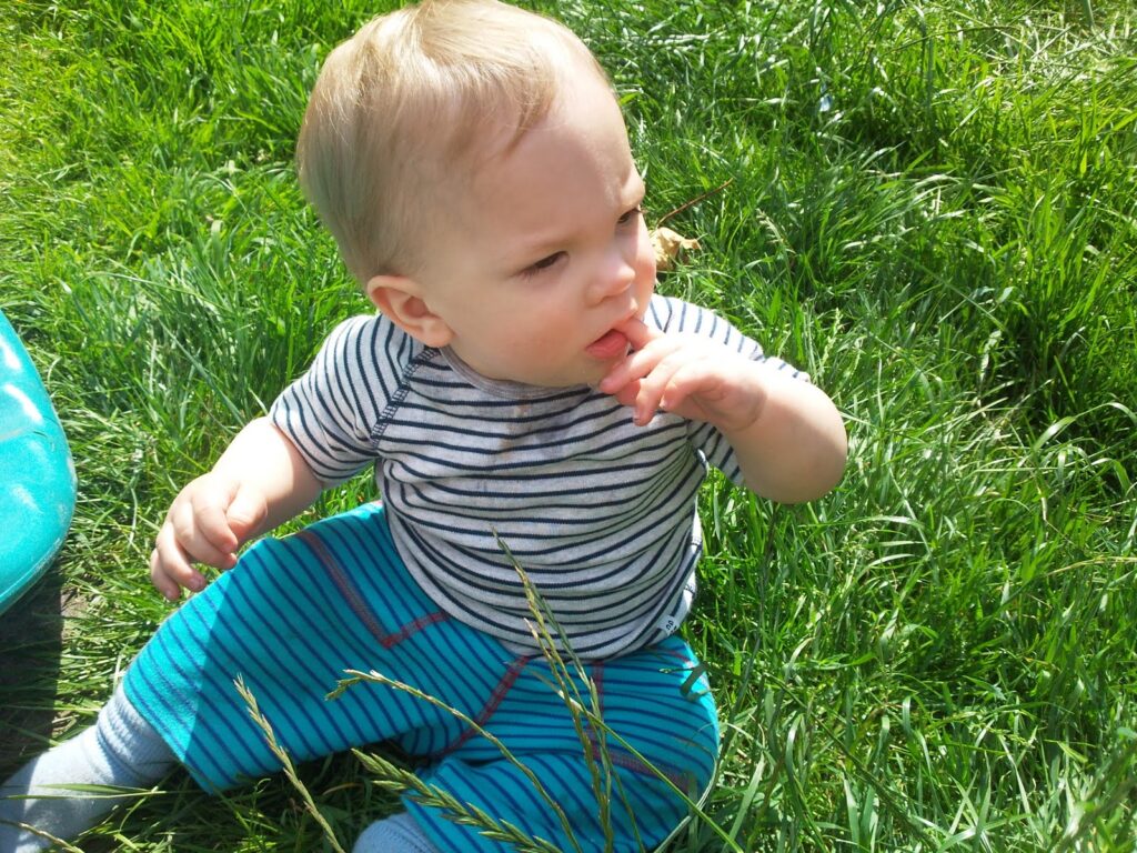 A baby with fair hair, wearing a striped grey and white shirt and blue striped trousers, sits in bright green grass. The baby touches their mouth with one hand and looks to the side, sunlight illuminating their face.