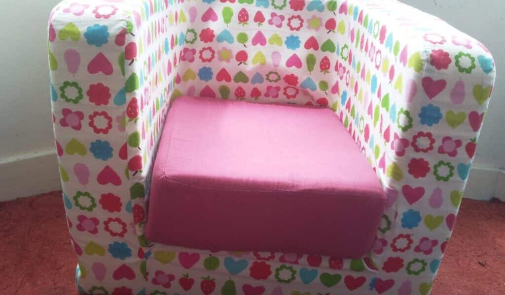 A small, boxy armchair for children covered in colourful patterns of hearts, flowers, and fruit shapes. The seat cushion is solid pink. The chair sits on a red carpet in front of a white wall with a narrow skirting board.